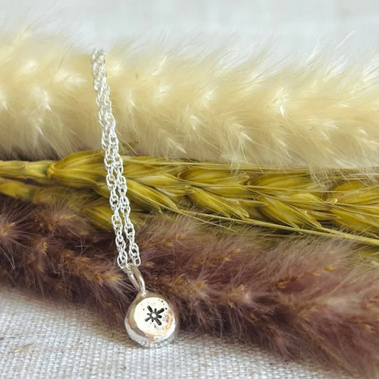 Silver recycled nugget pendant with star motif, on a silver chain placed on a sand coloured linen table cloth and a little bunch of dried foliage. 